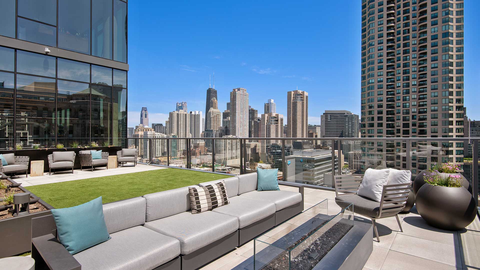 An outdoor couch and fireplace are in the foreground. Behind is a turf area for playing outdoor games and additional seating. Beyond the deck is the Chicago skyline on a clear day.