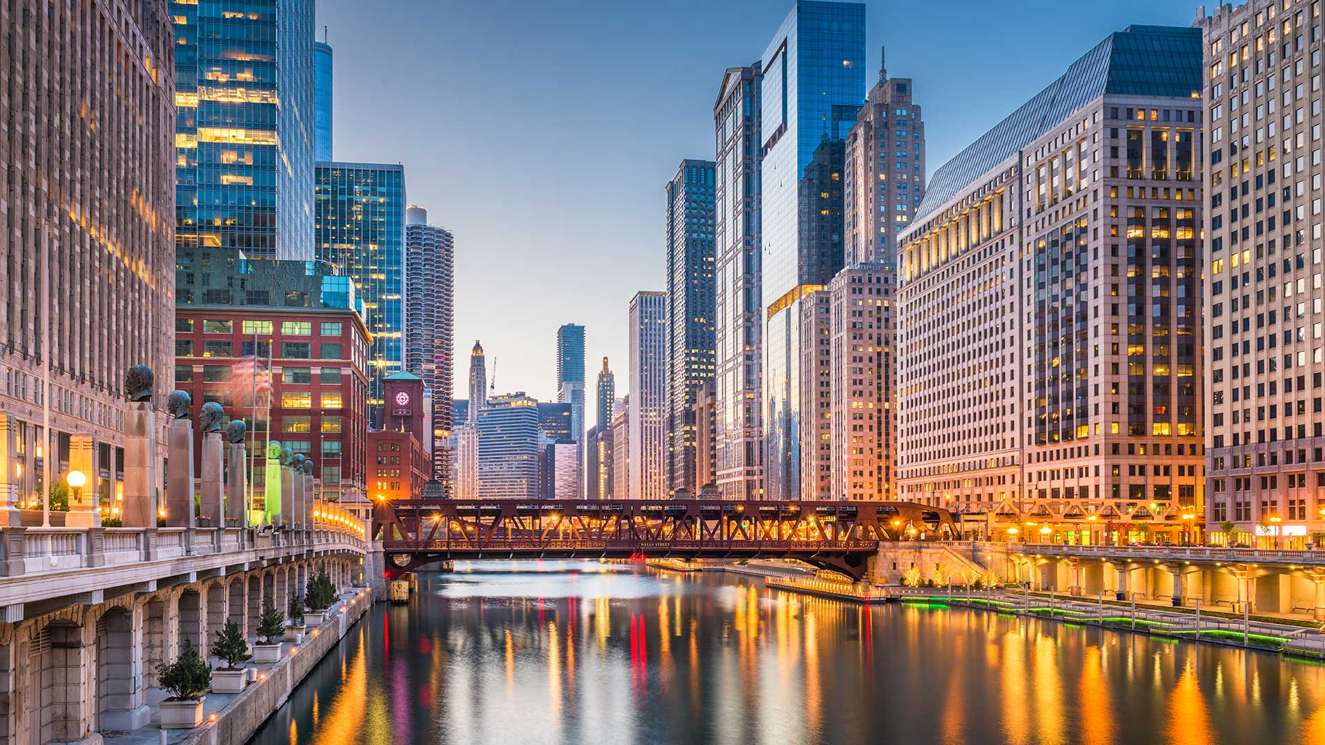 Looking down along the Chicago River at dusk. City lights are coming on, so the river reflects the many colors of those lights. Buildings rise all around the river on a clear evening.