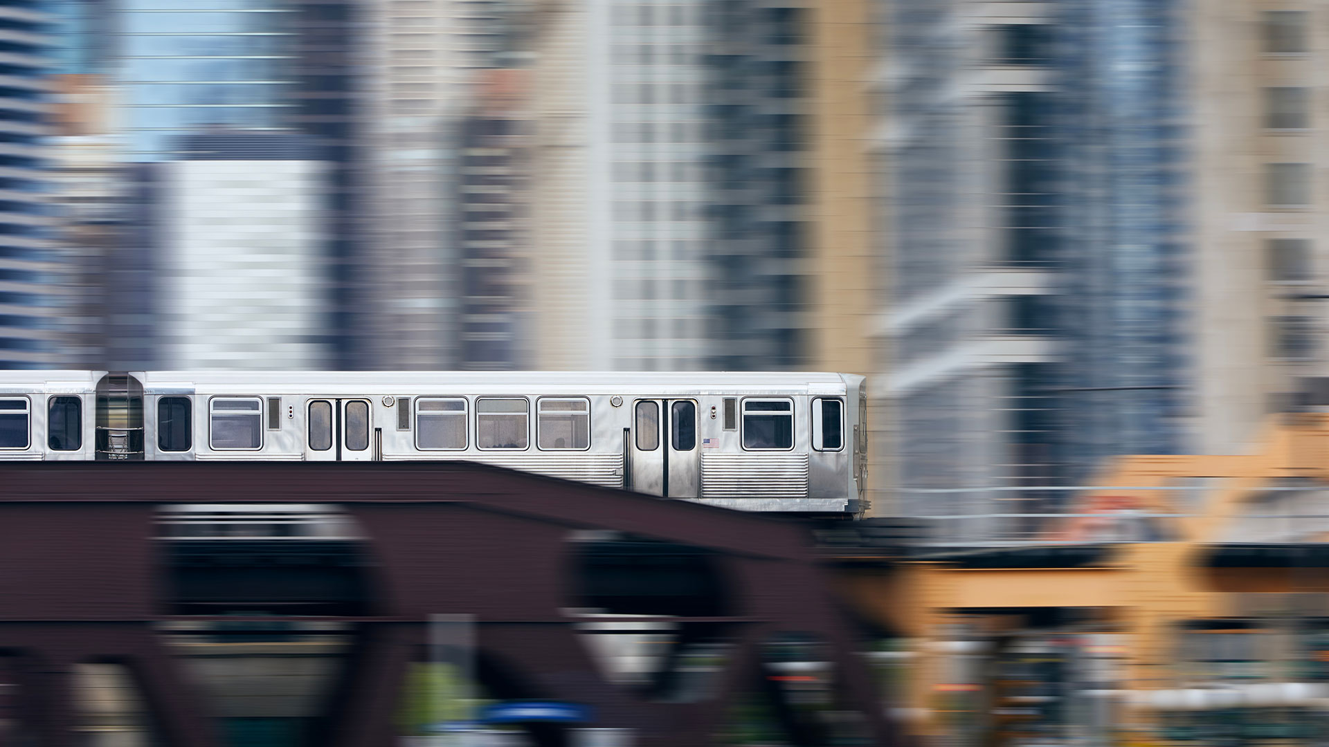 A silver CTA train seen in motion crossing a bridge. The rest of the image has a motion blur, but it appears there are city buildings zooming by.