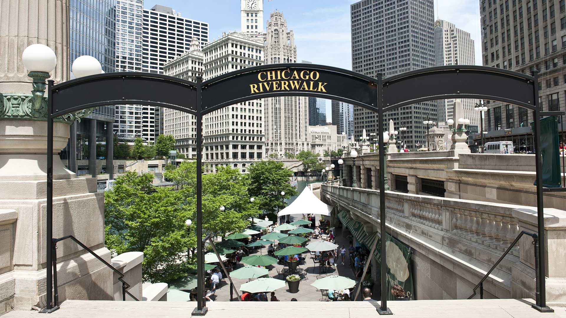Standing at the top of a staircase leading down to the Chicago Riverwalk. A sign that reads “Chicago Riverwalk” arches over the top of the stairs. There are café tables and open umbrellas seen below, as well as many trees. There are city building all around.