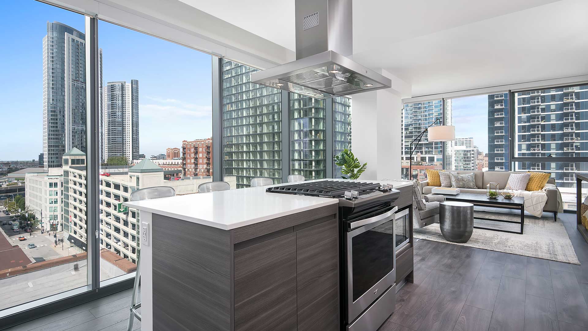 Looking across a kitchen island in a Hubbard221 residence. There is a stainless steel stove with range in the island, a stainless steel exhaust hood above. Beyond the kitchen to the right is a living room set. The walls are all floor-to-ceiling windows with city buildings seen outside.