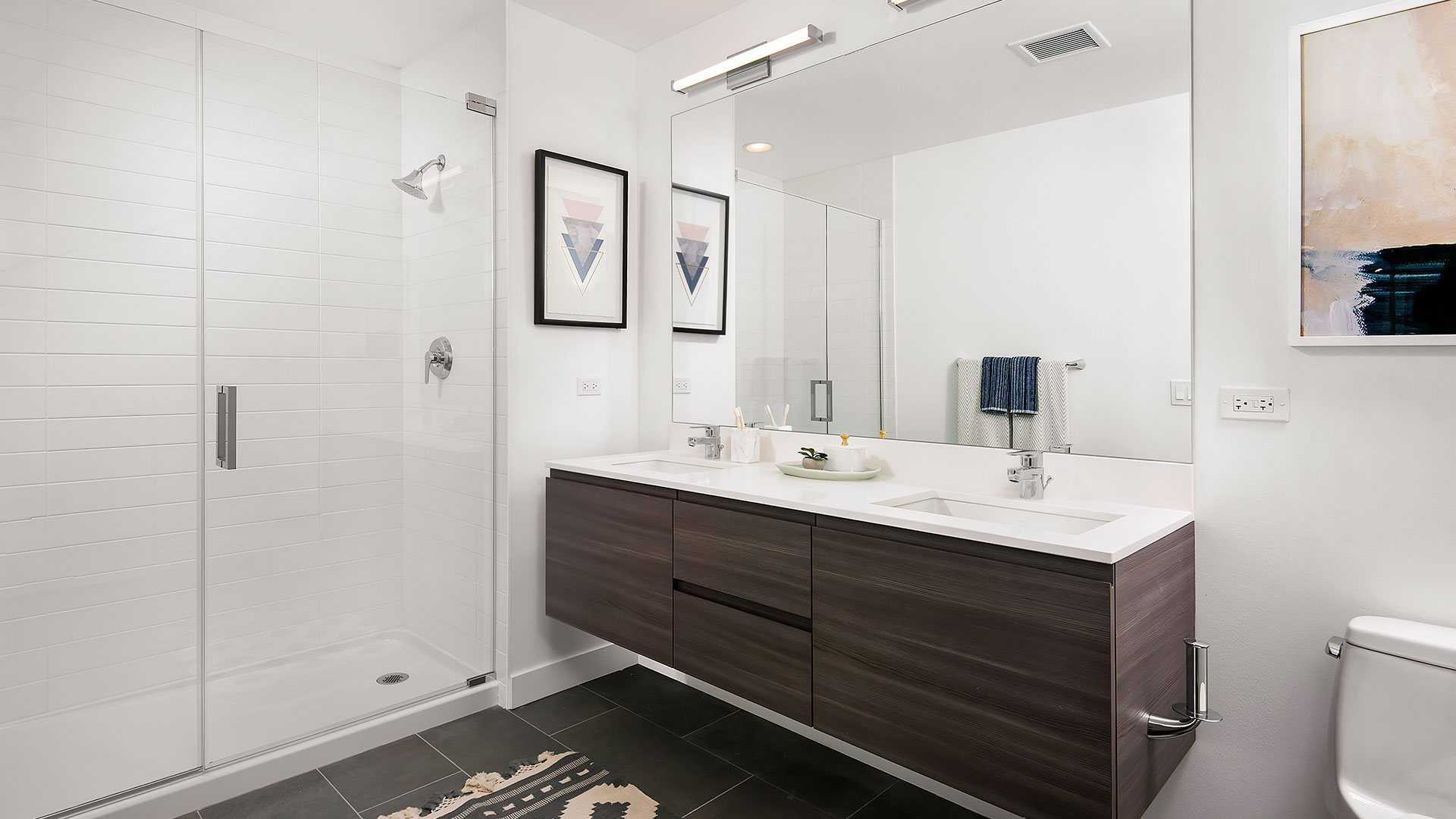 A bathroom in a Hubbard221 residence. A standing shower is on the left with a glass, frameless door. The double vanity is floating on the right with a full mirror mounted behind.