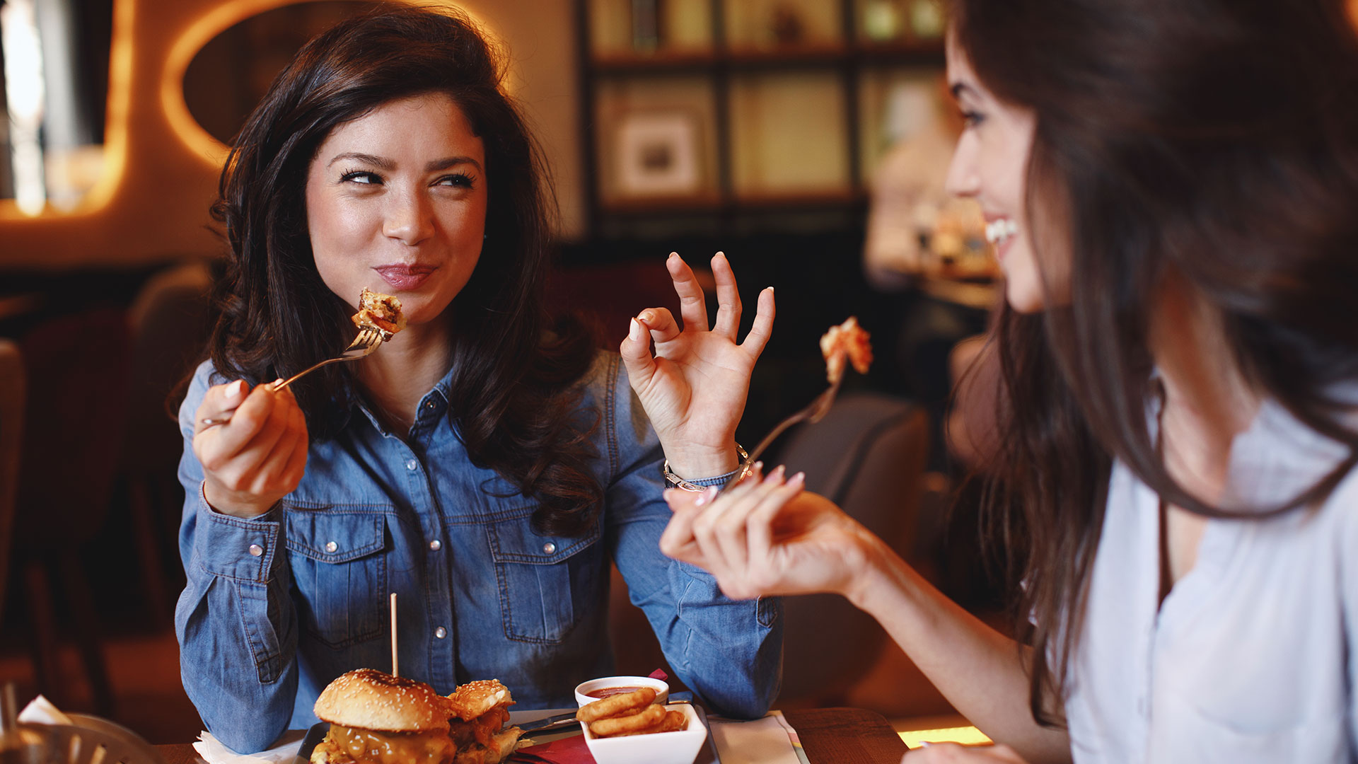 Two woman sharing a meal at a restaurant. The woman on the right is facing the camera holding up another bite to her mouth with one hand and gesturing that the food is good with her other. The woman on the left is also about to take another bite.